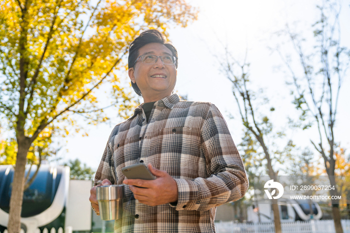 一个老年男人站在露营院子里拿着手机喝咖啡