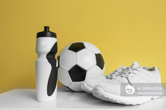 Bottle of water, soccer ball and sneakers on table near color wall