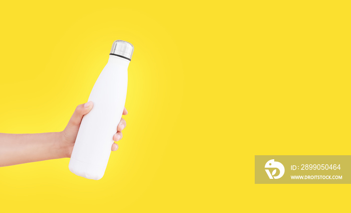 Close-up of female hand holding white reusable steel thermo water bottle isolated on background of yellow color with copy space.