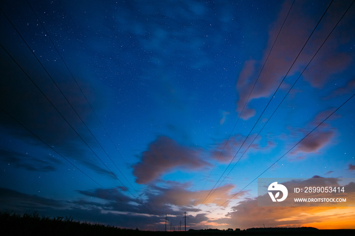 Night Starry Sky With Glowing Stars Above Countryside Landscape. Light Cloudiness Overcast Above Rural Field Meadow In Summer. Power Lines Above Rural Landscape.