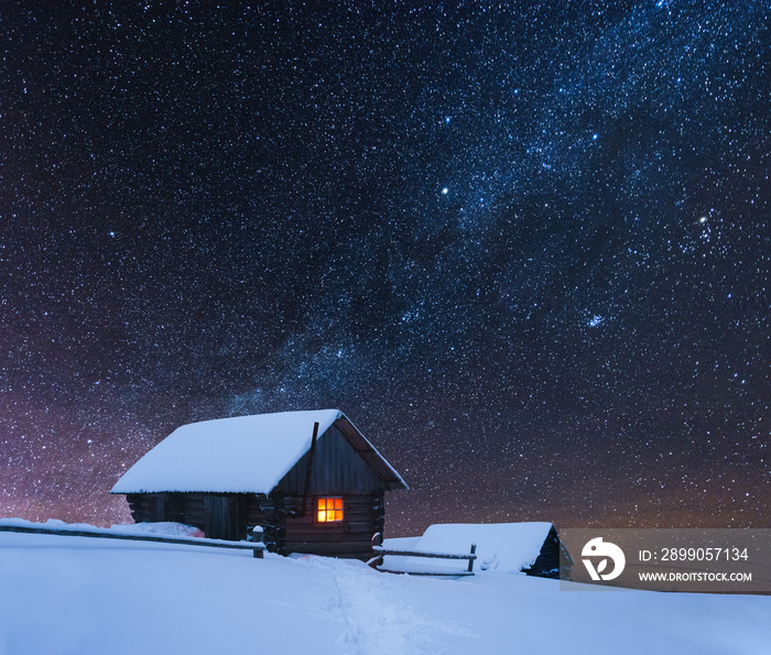 Dramatic wintry scene with snowy house with warm light in window. Fantastic night landscape glowing by milky way. Winter in Carpathian mountains, Ukraine, Europe