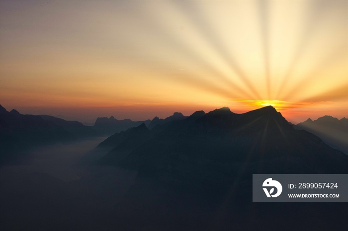 Sunbeams over Swiss mountain peaks at sunset time with valley and lake, colorful landscape picture