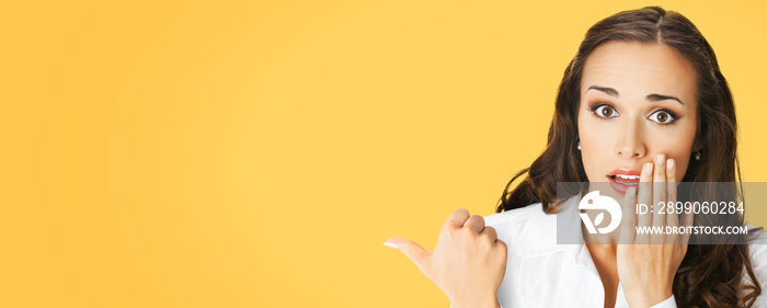 Portrait image of excited surprised, shocked astonished businesswoman pointing away side with finger. Business woman covering open mouth by her hand. Yellow color background. Wide banner composition.