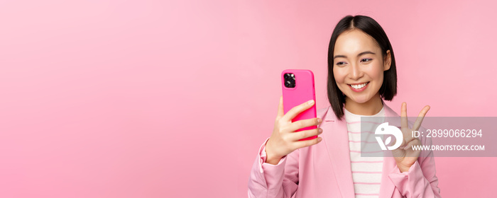 Stylish asian businesswoman, girl in suit taking selfie on smartphone, video chat with mobile phone app, posing against pink studio background