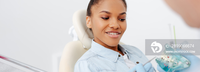 panoramic shot of happy african american woman in braces near dentist holding teeth model