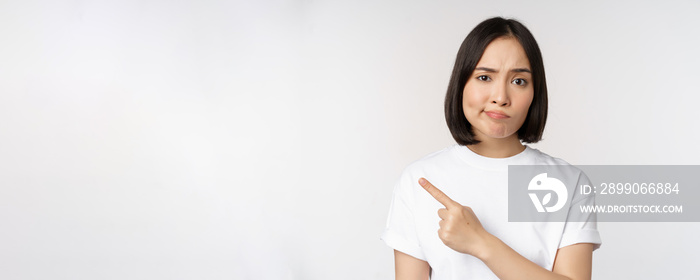 Skeptical asian girl in white t-shirt, pointing at product or logo with disappointed grimace, dislike and complain at smth, standing over white background