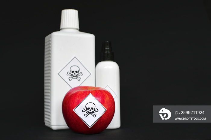 Concept of pesticide residues in agricultural food products dangerous to humans, showing a red apple and white bottles with poison symbol sticker on black background