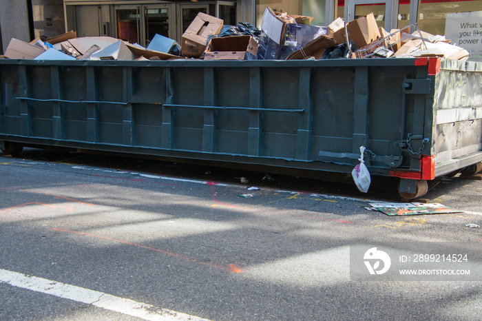 Long overflowing metal dumpster on a asphalt street in front of businesses