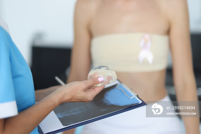 Doctor’s hands are holding breast implant in front of clipboard with patient in background. Mammaplasty breast correction concept.