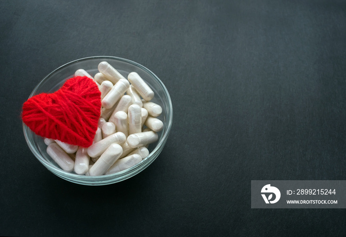 White veg capsules and red thread heart in glass bowl on black background. Anticoagulant, Blood Thinners. Cardiac Medications, pills for the heart. Treat Heart disease Failure