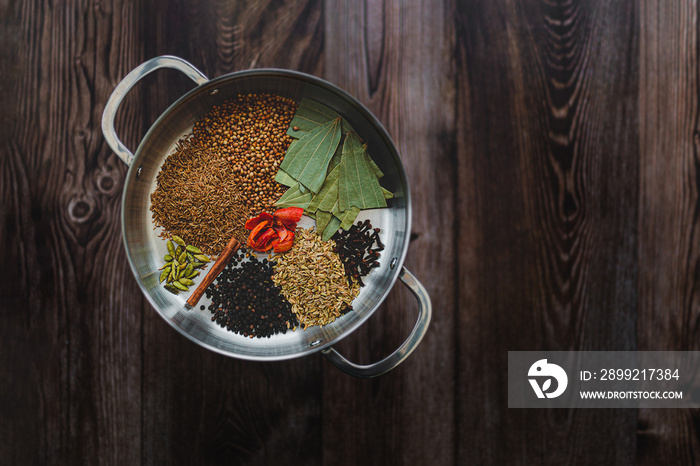 Indian curry spices arranged in a metal pan and placed on left side of frame. Dish is on dark wooden table with texture and natural age. Dark mood food photo with copy space available.