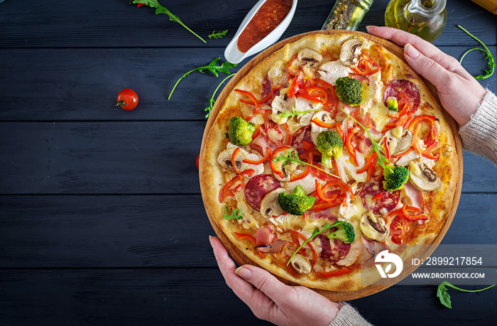 Fresh Italian pizza with chicken fillet, mushrooms, ham, salami, tomatoes, cheese on a black background in the hands. Italian food. Top view