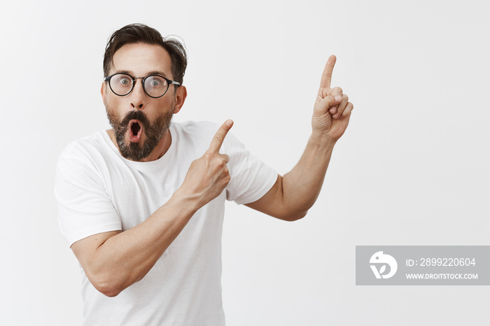Studio shot of shocked and surprised impressed man with beard and moustache in trendy black glasses, pointing at upper right corner with both hands, dropping jaw and staring shook at camera