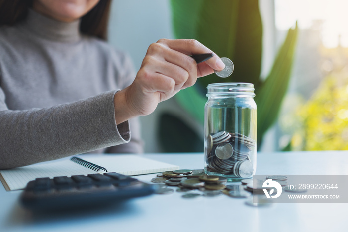 Closeup image of a woman putting coins in a glass jar with calculator on the table for saving money and financial concept