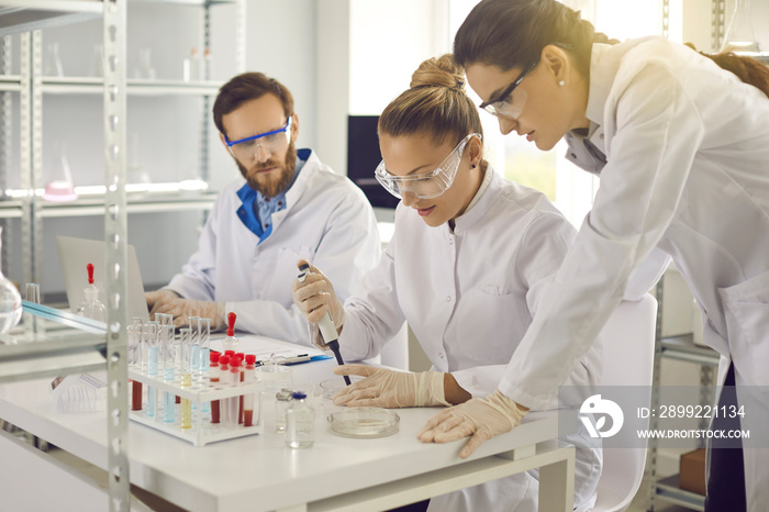 Group of scientists working in science laboratory. Pharma chemists or biotech company employees in lab coats and goggles preparing to conduct experiment and transferring sample liquid on Petri dish