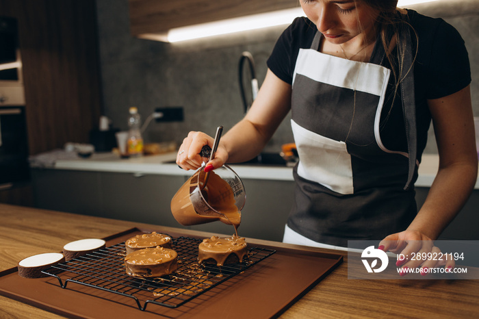 Smiling female confectioner pouring liquid chocolate gommage on biscuit cake. Woman chef in uniform at assembling mousse dessert process