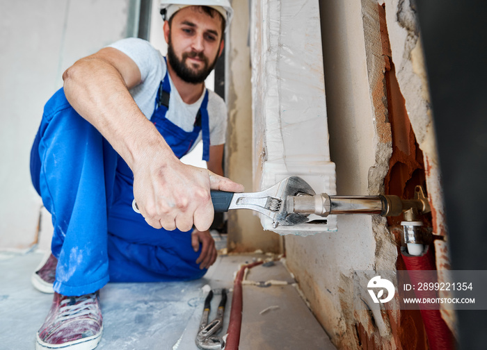 Bearded young man in work overalls using plumber wrench while installing heating radiator in room. Male plumber in safety helmet installing heating system in apartment. Focus on hand with instrument.