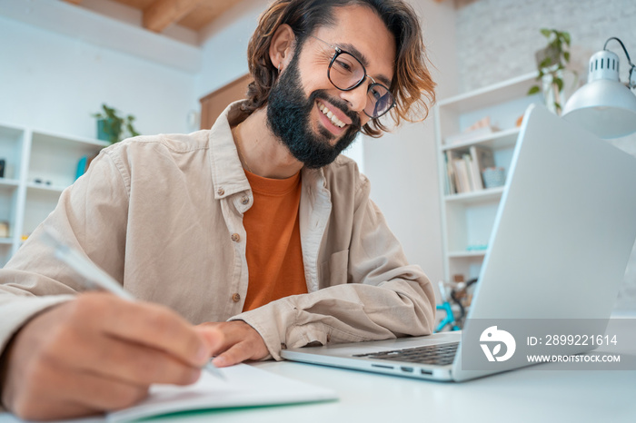 Happy young creative entrepreneur laughing smiling writes in a notebook next to laptop in a modern workspace office at home. Freelance guy starting small business. High quality photo