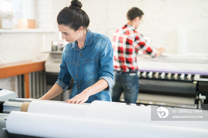 Serious concentrated woman in denim shirt standing at large format printer and choosing program while working in printing office