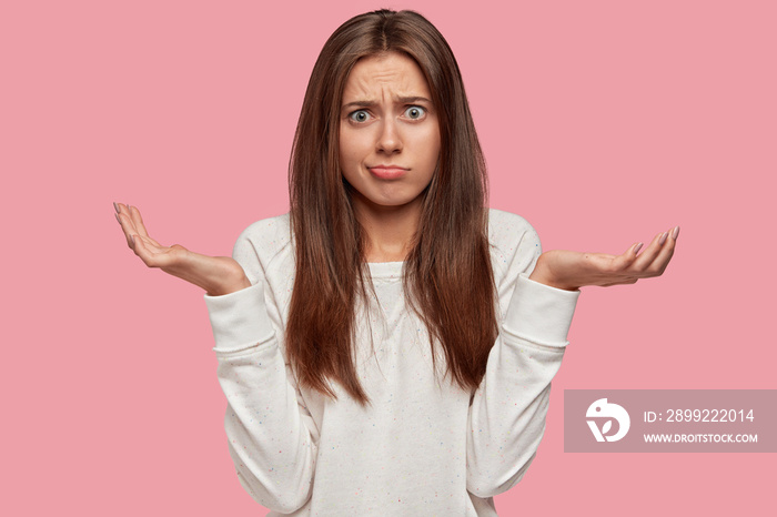 Indignant lovely woman shruggs shoulders, has hesitant expression, dressed in casual white sweater, expresses apathy, isolated over pink background, feels clueless about her future. Puzzlement concept