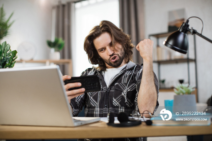 Happy caucasian man student in casual wear sitting at table with modern laptop and keeping arm raised. Concept of people, technology and distance learning and success.