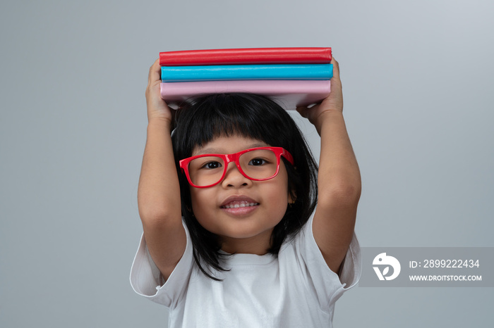 Funny and Happy Asian little preschool girl wearing red glasses holding a green book on the head, on white isolated background. Concept of school kid and education in elementary and preschool