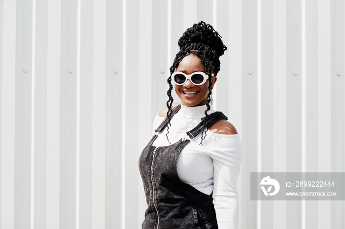 African woman in denim overalls skirt, white sunglasses posed against white steel wall.