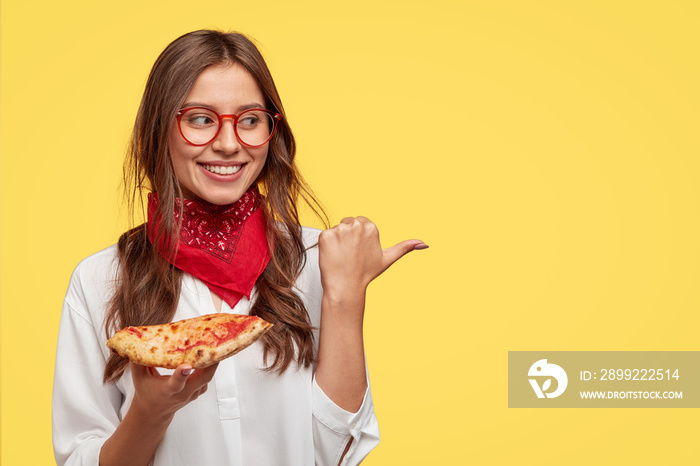 Isolated shot of attractive smiling girl shows direction to pizzeria, eats tasty pizza with cheese and tomatoes, points with thumb at copy space against yellow background. Woman has snack indoor