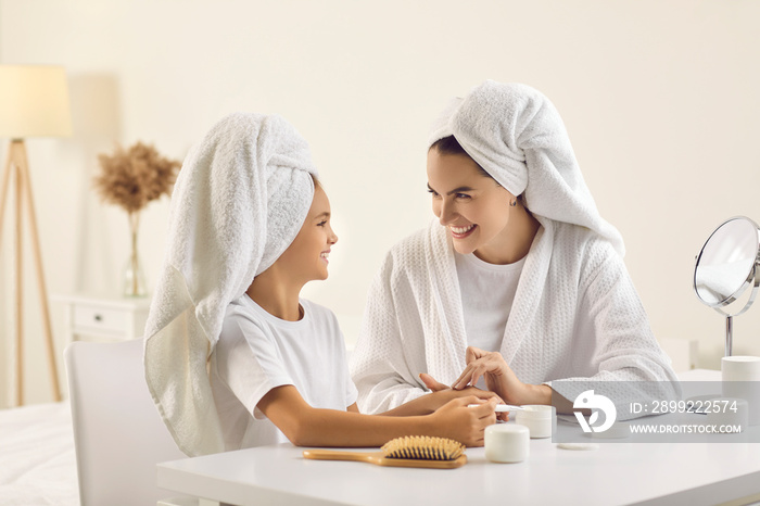 Young mom showing child daily skin care routine. Happy mother and pretty little daughter in white bath towels sitting at beauty table in bedroom, enjoying hand and face masks and looking at each other