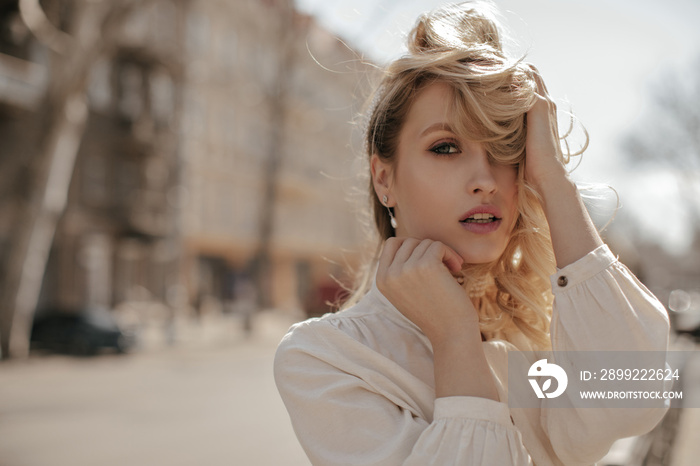 Grey-eyed curly blonde woman in white stylish blouse touches hair and looks into camera. Pretty lady walks outside.