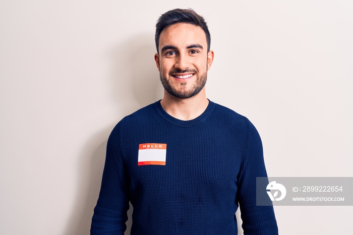 Young handsome man with beard wearing identification sticker over isolated white background looking positive and happy standing and smiling with a confident smile showing teeth