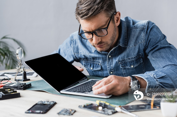 Focused repairman with broken key looking at keyboard of damaged laptop at workplace on blurred foreground