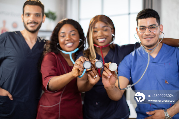 Blur front view of four young diverse multiethnic medical practitioners, standing together in light hospital room and demonstrating stethoscopes to camera. Focue on stethoscope heads