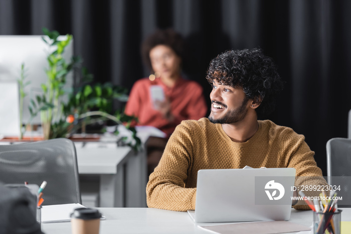 happy indian advertising manager looking away near laptop and blurred african american colleague.