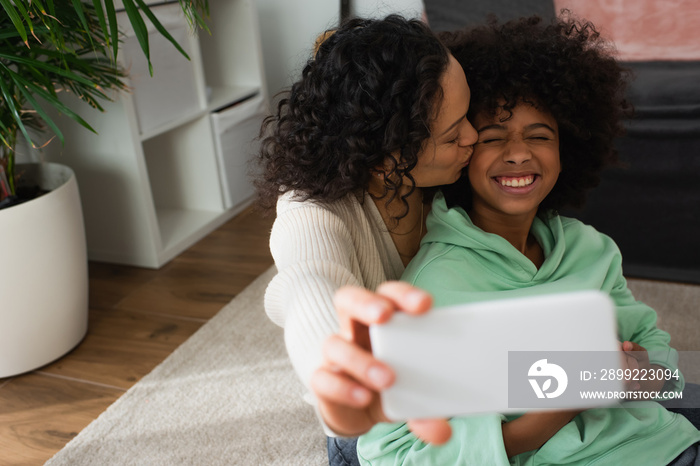 curly african american mother kissing cheek of daughter and taking selfie on smartphone.