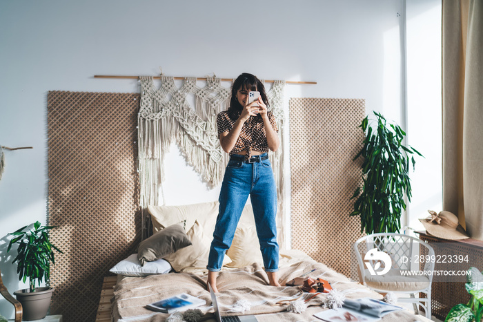 Cheerful woman taking photo while standing on bed in boho room