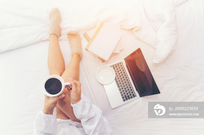 Close up legs women on white bed in the bedroom. Women working on laptop and drinking coffee in morning relax mood in winter season. Lifestyle Concept.