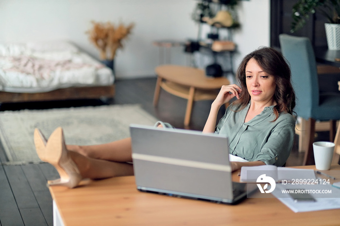 Attractive charming successful manager is having a rest, she is watching educational video on the laptop and put her feet on the table. Freelancer using laptop and the Internet.