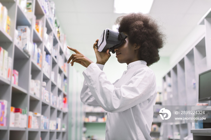 Side view of young professional African woman pharmacist, wearing virtual reality headset, working in modern drugstore and looking at shelves with medicines and gesturing