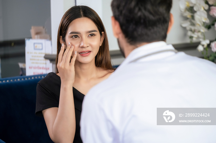 Woman discuss with confident doctor who wear lab coat and sitting on sofa at the modern beauty clinic. Beautiful female touch her face and need to do plastic surgery. Beauty and service concept.