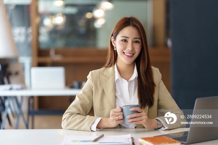 Beautiful Asian business woman holding a coffee mug and laptop at the office. Looking at the camera.