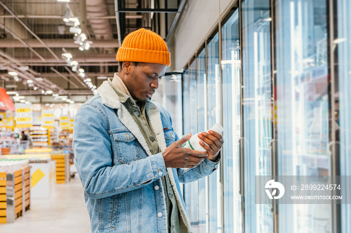 Positive African-American guy in denim jacket and orange hat takes milk bottle from fridge case in modern supermarket side view