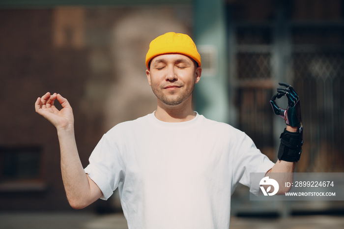 Disabled european man with artificial limb of hand meditating outdoors. Handsome young guy wears t-shirt and hat enjoys sunny day. Concept of mental health. Modern medical technology today