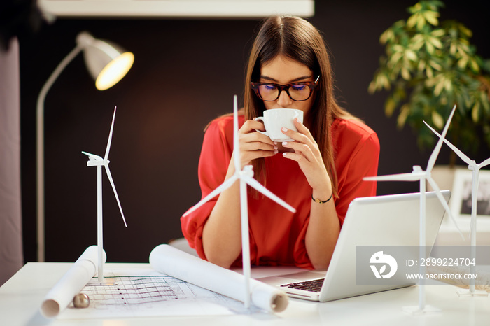 Beautiful caucasian businesswoman dressed in red blouse sitting in modern office and driking coffee. On table are, blueprints, laptop and windmill models. Sustainable development concept.