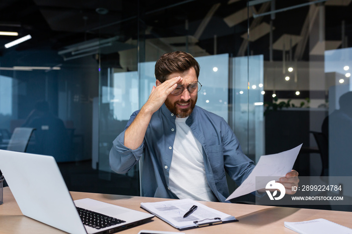 Upset businessman behind paper work inside modern office, mature man with beard reading financial reports and account documents unhappy with results and disappointed with achievements.