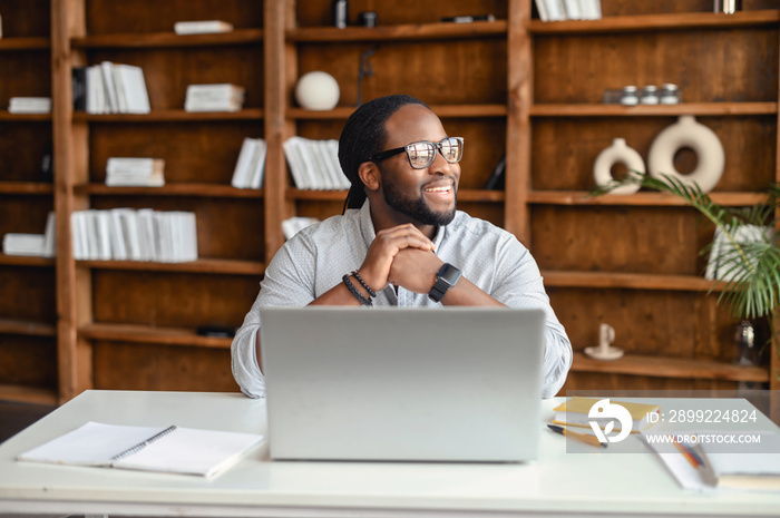 Happy thoughtful young African American man in glasses sitting at desk in the office, looking away, taking break from working on laptop, thinking of new ideas, smiling and planning weekend or vacation