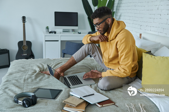 Confident African man using laptop while sitting on the bed at home