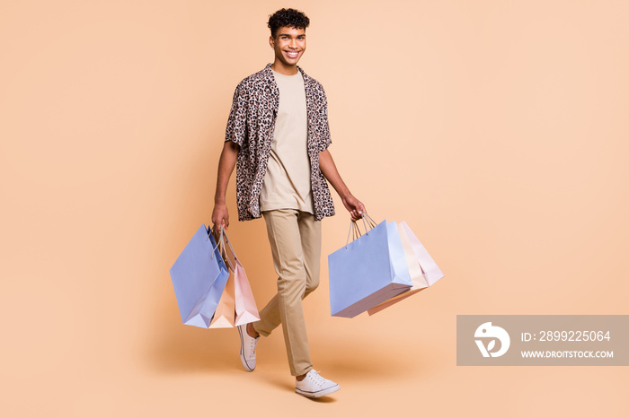 Full length portrait of positive dark skin man carry bags look camera toothy smile isolated on beige color background