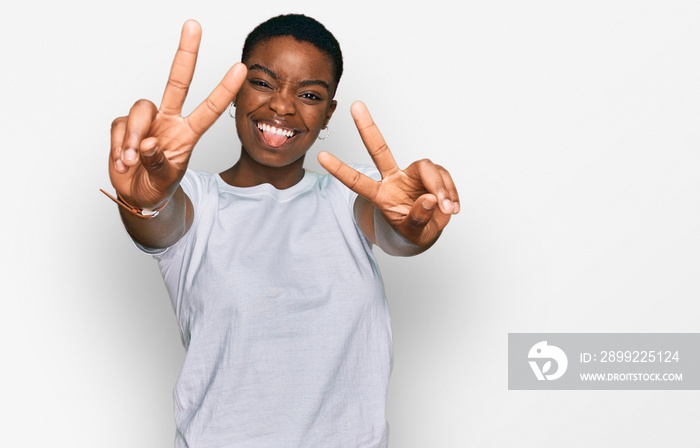 Young african american woman wearing casual white t shirt smiling with tongue out showing fingers of both hands doing victory sign. number two.