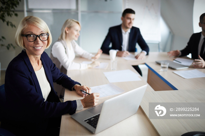 Successful mature businesswoman sitting by table in front of laptop and camera with her subordinates on background